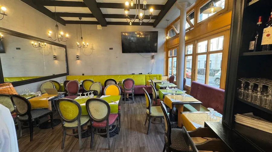 La salle — banquettes et chaises médaillon en velours, poutres noires, grandes fenêtres sur la rue