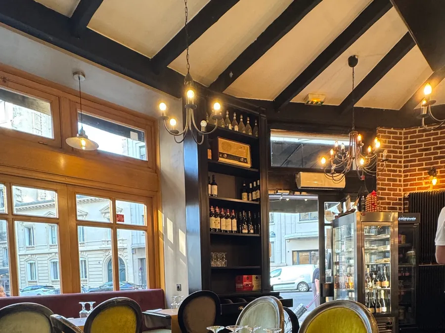 La salle — poutres noires, lustres en fer, étagère à vins et mur de briques, fenêtres sur la rue